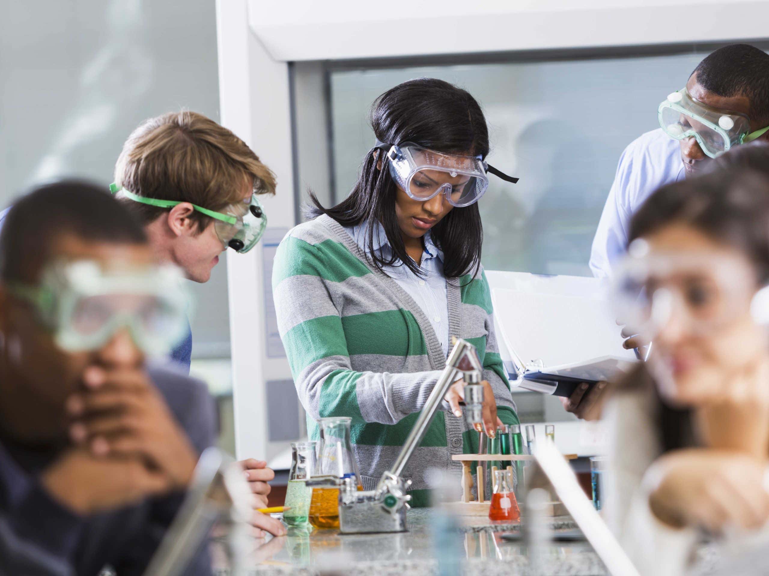 Multi-ethnic group of students with instructor in chemistry lab, wearing safety goggles, working with chemicals in test tubes and beakers. Selective focus.