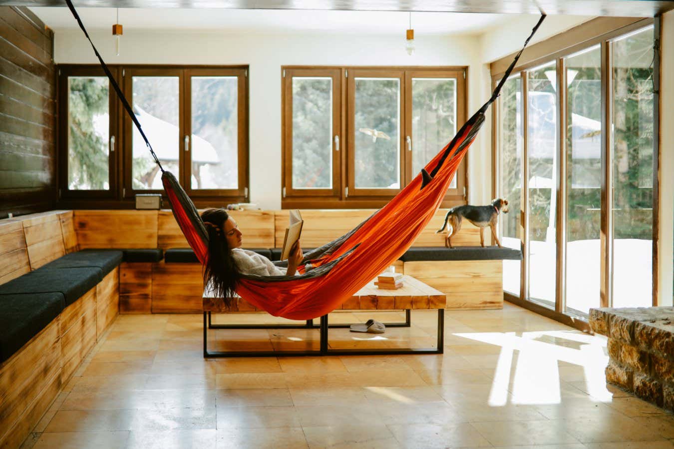 Woman in her late 30s reading a book in the mountain cabin, relaxing in the hammock next to the fireplace.